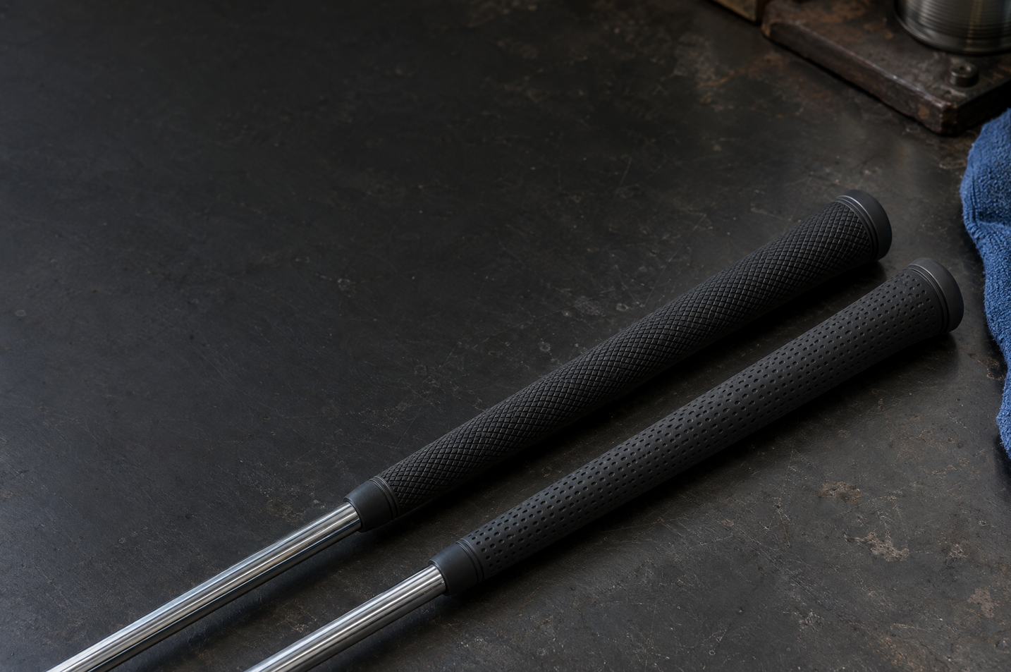 Two black textured golf grips on steel shafts lying on a dark workbench beside a blue cloth.
